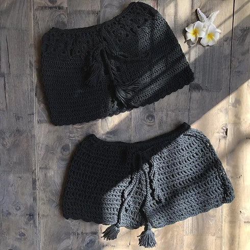 Black crochet beach shorts with tassel drawstrings on wood, tropical flowers nearby