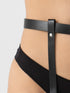 Close-up of a model's lower torso wearing a black faux-leather garter belt with a vertical strap and silver-tone rivet, paired with black underwear against a light gray background.