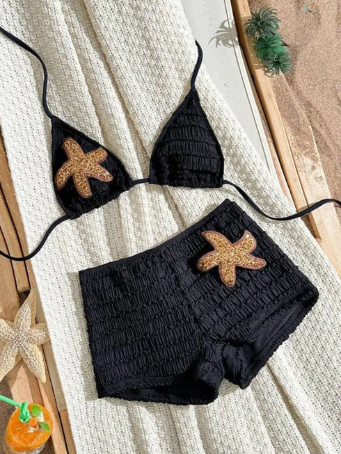 Black textured bikini set with starfish appliqués, laid flat on a cream knit blanket with beach-themed props.