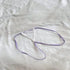 A delicate lavender and white strappy jewelry accessory, possibly a small necklace or pendant, laid flat on a textured white fabric background.