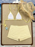 A two-piece bikini set displayed on a beige board, featuring a white triangle bikini top with yellow trim and matching yellow ruffled shorts. A straw hat, starfish charms, and a beaded necklace are styled around it against a beach backdrop.