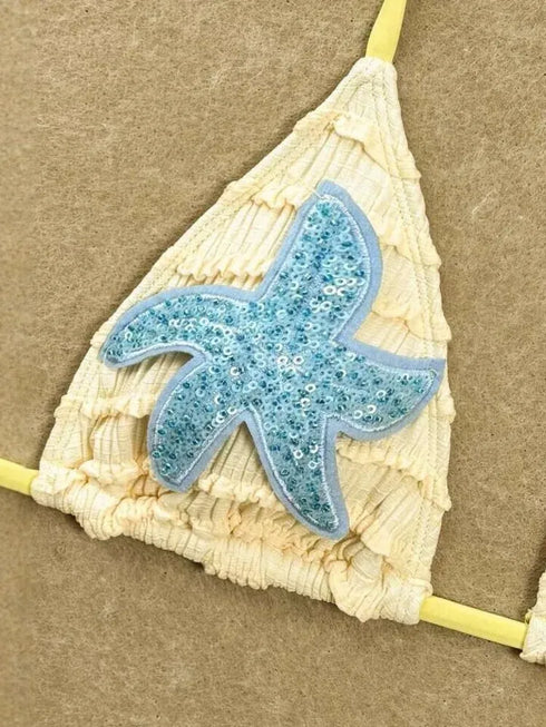 A close-up of a bikini top strap with a cream-colored textured fabric and a light blue sequined starfish applique on a tan background.
