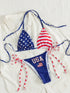 Patriotic USA-themed string bikini laid flat on a white button-down shirt. The bikini top has one star-spangled blue cup and one red-and-white striped cup, with blue string ties.
